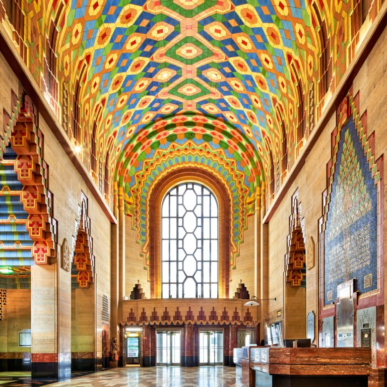 A grand hall with a high, colorful geometric ceiling, large arched window, detailed walls, and a reception desk in an Art Deco-style building.