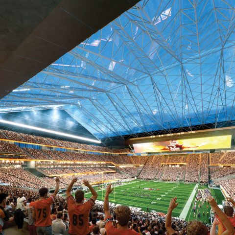 Large stadium filled with cheering fans watching a football game; the field is green and the retractable roof is partly open, revealing a blue sky above.