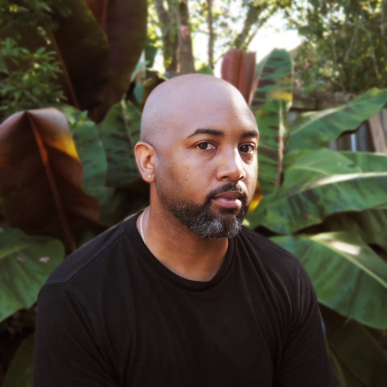 Bald man with a short beard and black shirt sits in front of large leafy green plants outdoors, looking at the camera.