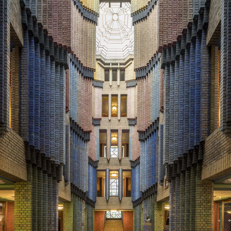 Symmetrical brick atrium with multicolored columns, geometric floor patterns, and a glass ceiling letting in natural light.