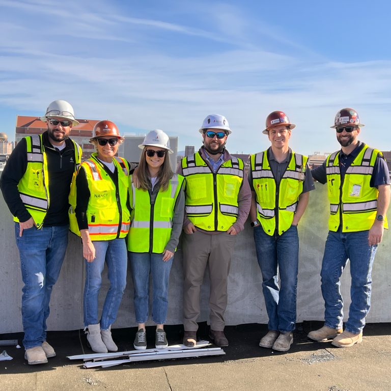 Six people wearing safety vests and hard hats stand side by side on a rooftop with a cityscape in the background.