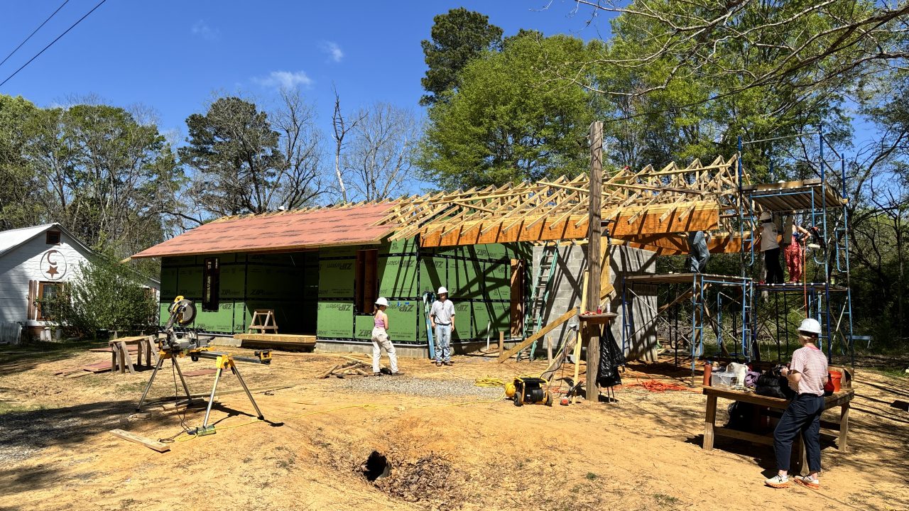 Five people work on a green house under construction, with exposed roof framing and scaffolding, surrounded by tools on a sunny day.