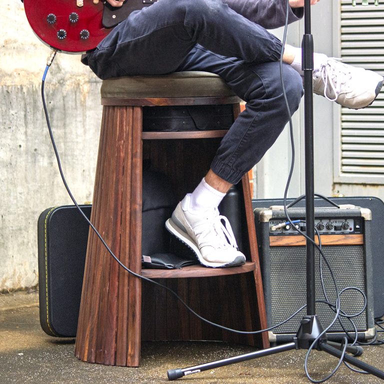 Person sitting on a wooden stool playing an electric guitar, with a guitar amplifier and cables on the ground nearby.