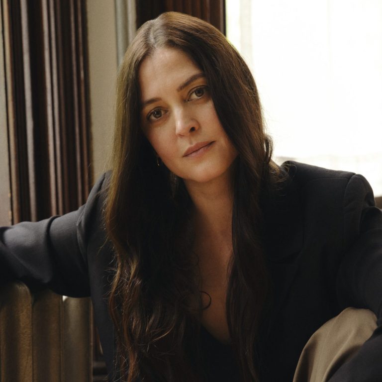 A woman with long brown hair sits by a radiator, wearing a black blazer and tan pants, with one arm resting on the radiator and a calm expression on her face.
