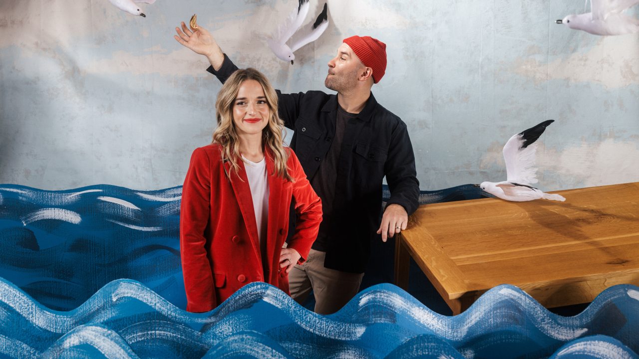 A woman in a red jacket stands in front of a man feeding seagulls by a wooden table, with blue wave decorations and a painted sky background.