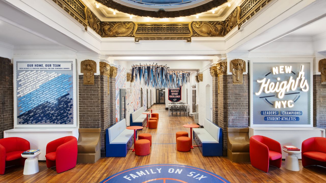 Colorful lounge area with red chairs, blue booths, basketball court floor design, and NYC-themed wall art under a skylight and ornate ceiling.