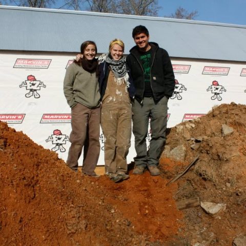 Three people in work clothes stand together smiling between two mounds of dirt in front of a building with Marvins logos on a banner.