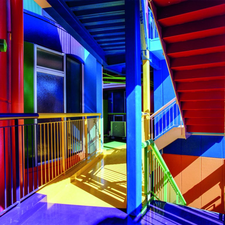 A brightly colored stairwell and hallway with red stairs, blue beams, yellow floor, and multicolored walls, lit by natural daylight.