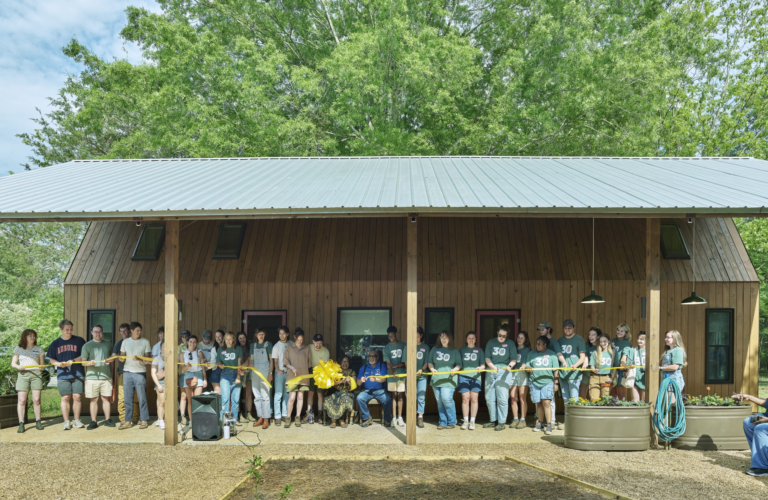 A group of people stand in front of a wooden building, holding a green ribbon stretched across the porch for a ribbon-cutting ceremony.
