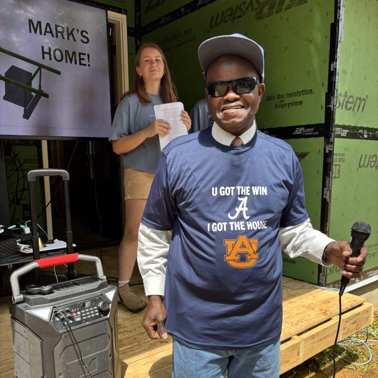 A smiling man holds a microphone and wears an Auburn shirt; a woman stands behind him on a porch with a sign that says MARKS HOME! and a speaker beside them.