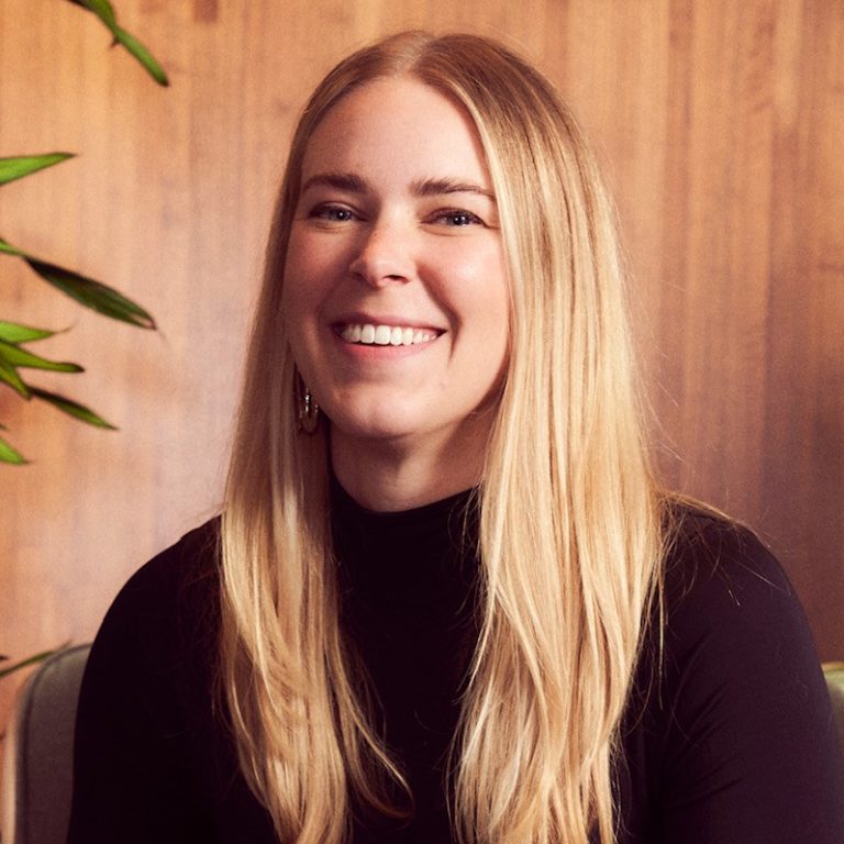 A woman with long blonde hair sits on a chair, smiling, wearing a black top, blue jeans, and cream boots. She is surrounded by potted green plants against a wooden wall.