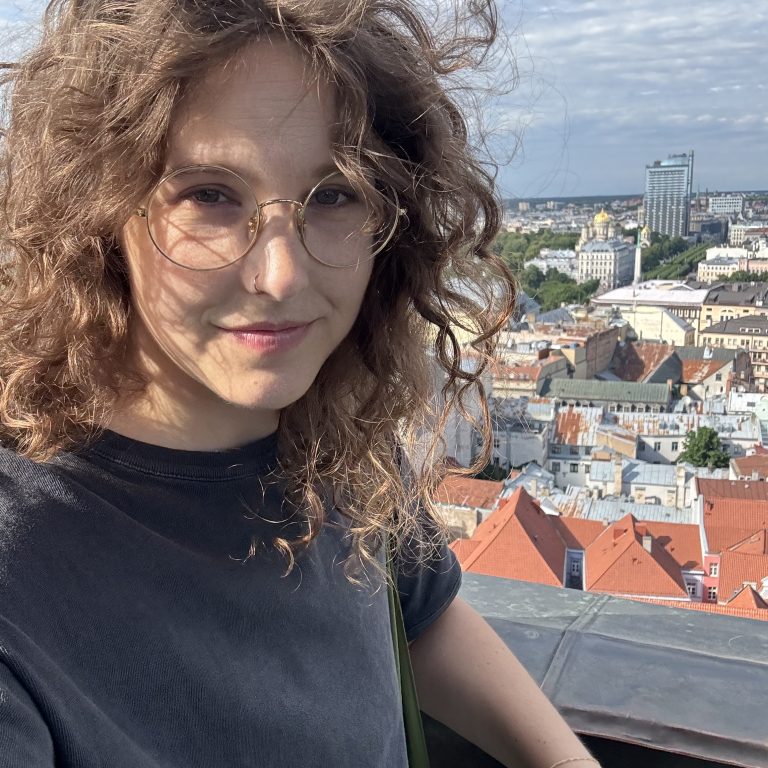 A person with curly hair and round glasses posing for a selfie overlooking a city with red-roofed buildings.