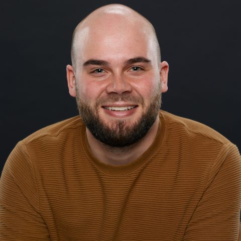 A smiling man with a shaved head and a beard wearing a brown long-sleeved shirt against a black background.